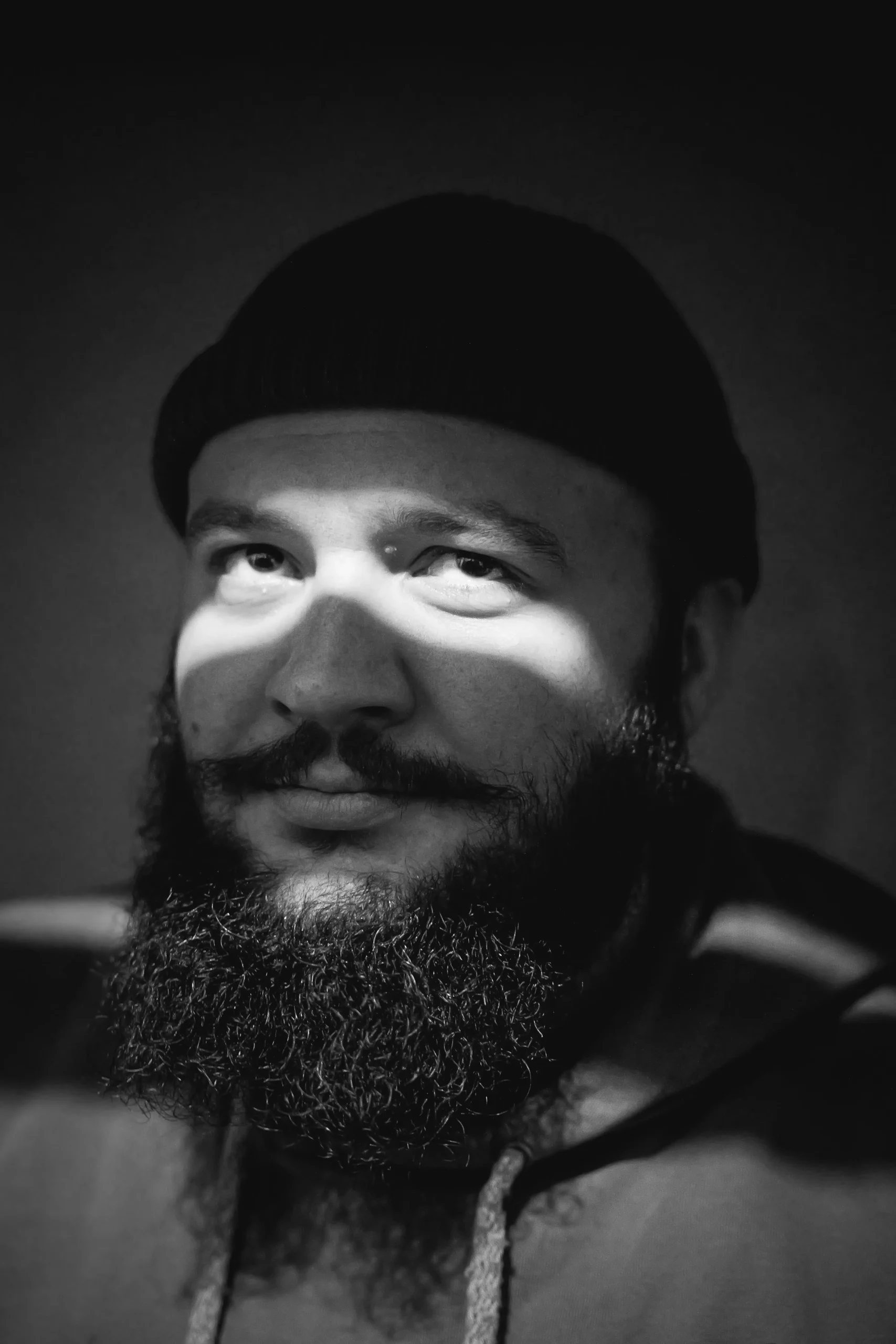 High-contrast black and white portrait of a bearded man with a dramatic shadow cast across his eyes.
