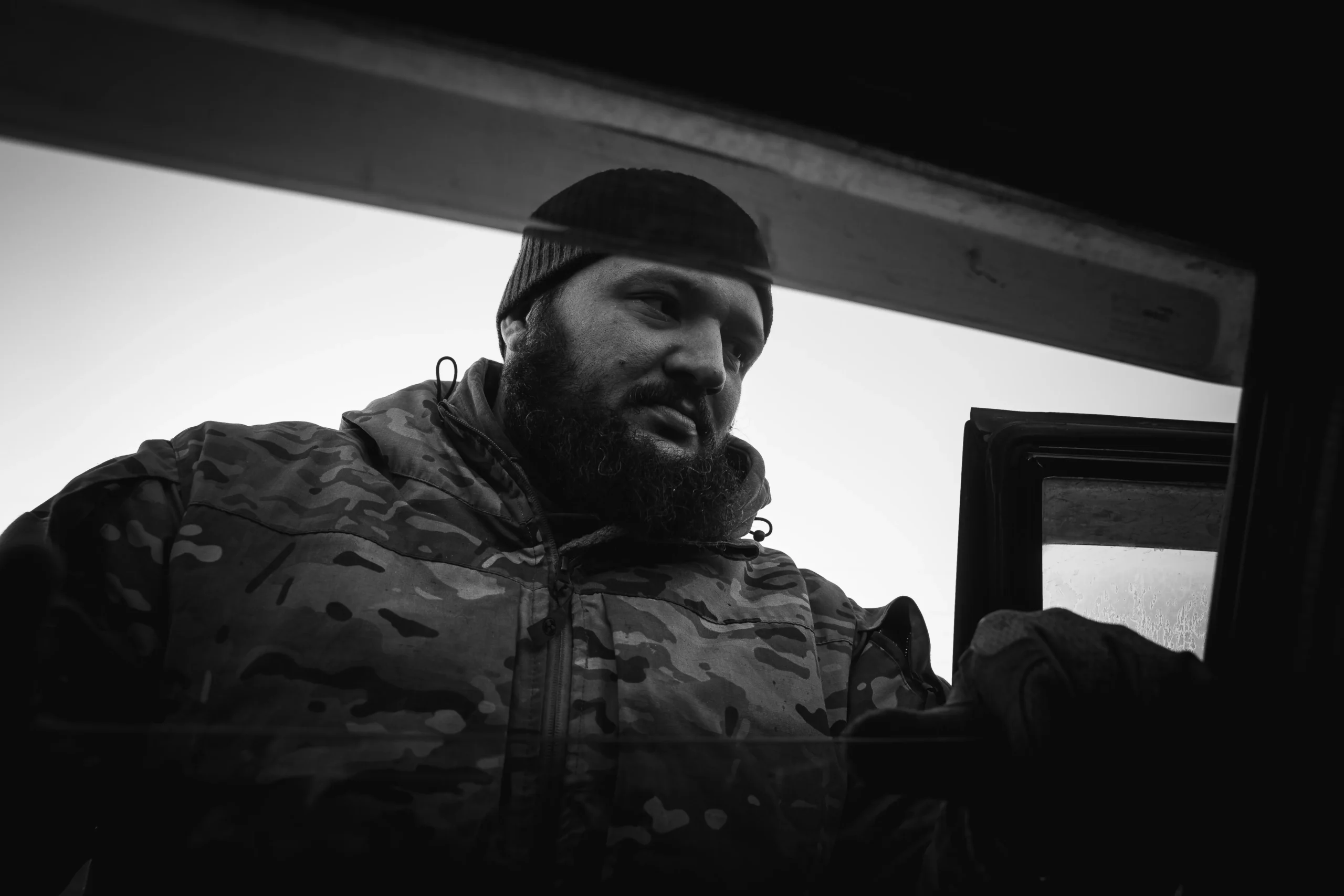 A bearded soldier in a camouflage jacket seen through the glass of a vehicle window, looking contemplative.