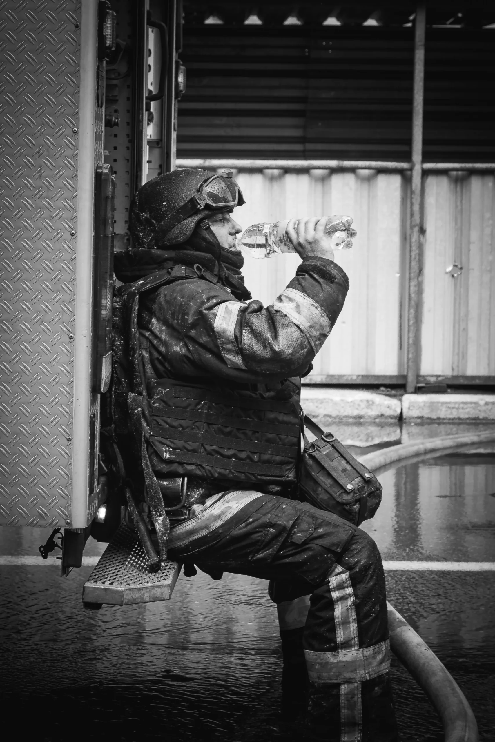 Black and white shot of an exhausted firefighter sitting on the back of a truck, drinking from a water bottle.