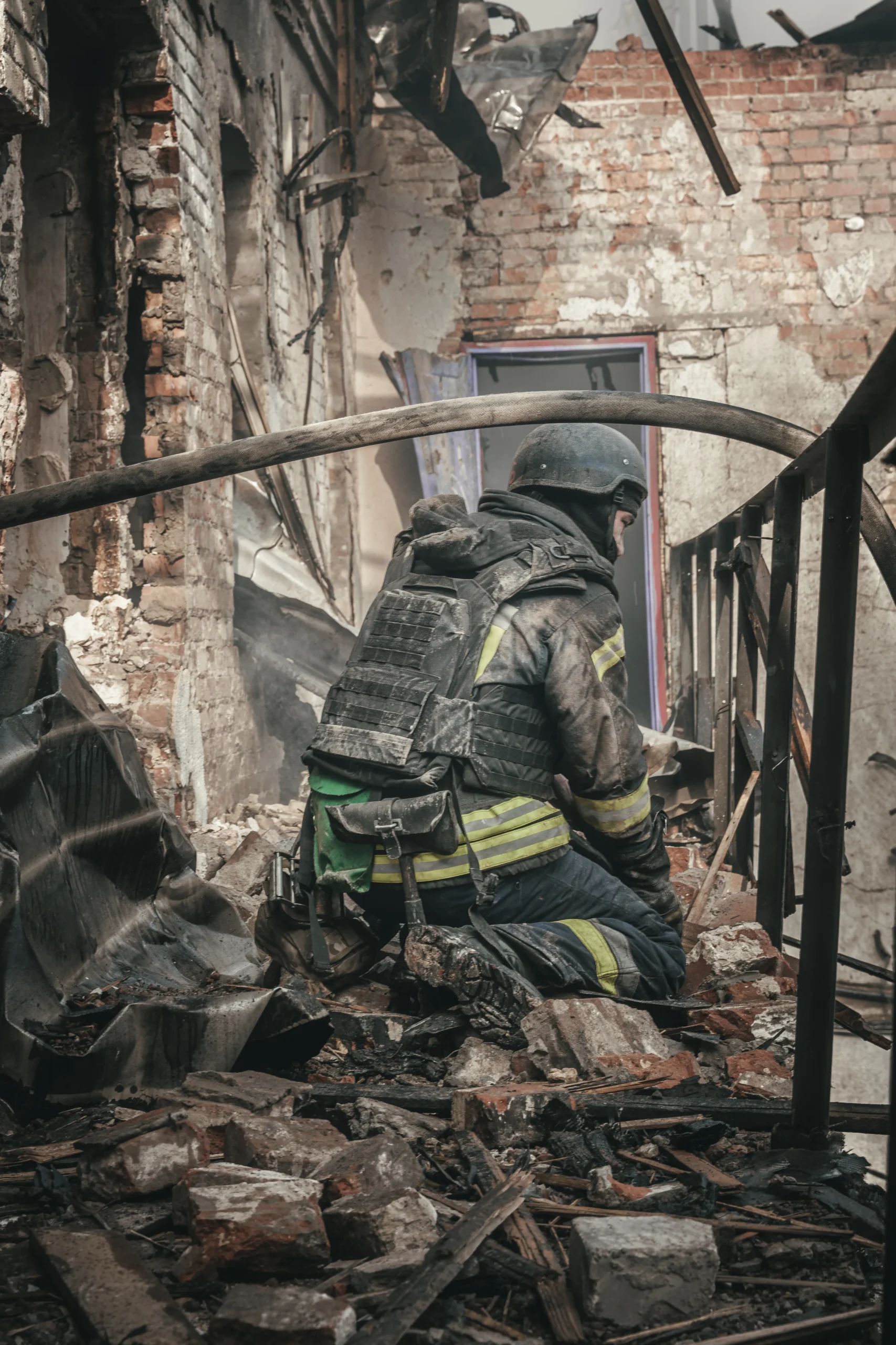 A firefighter in full protective gear and helmet kneeling amidst the brick rubble of a destroyed building.