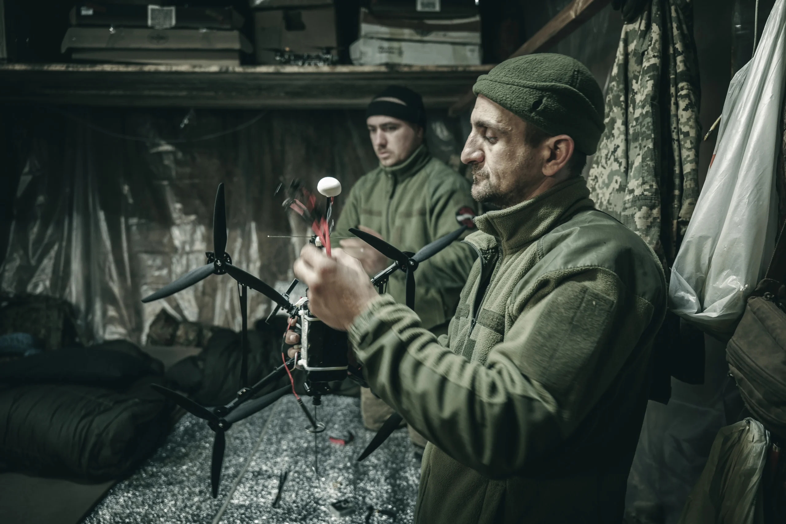 A military technician in a green fleece jacket working on a large black multi-rotor drone inside a dimly lit workshop.