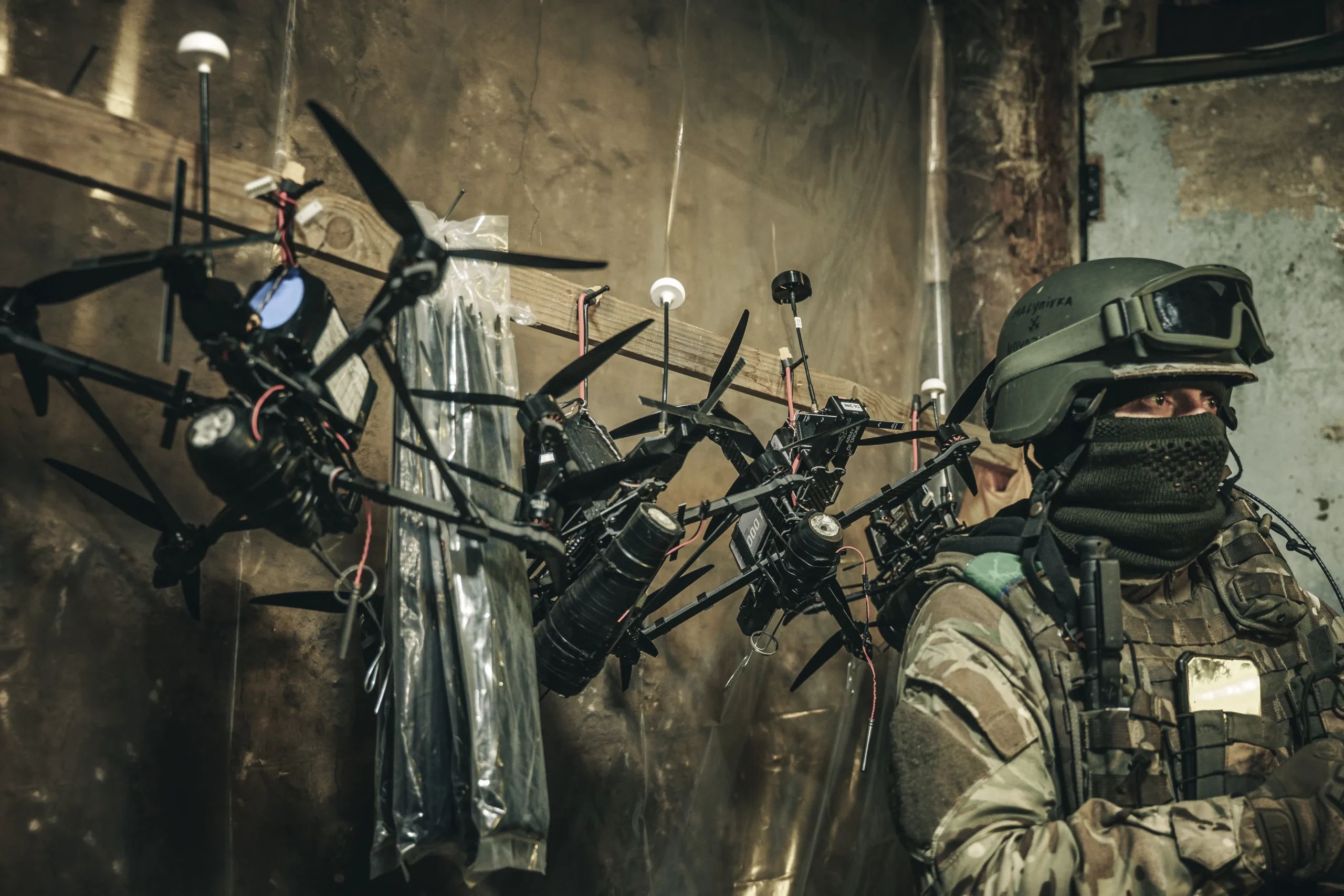Several FPV kamikaze drones hanging from a wooden rack inside a military shelter, with a soldier in the background.