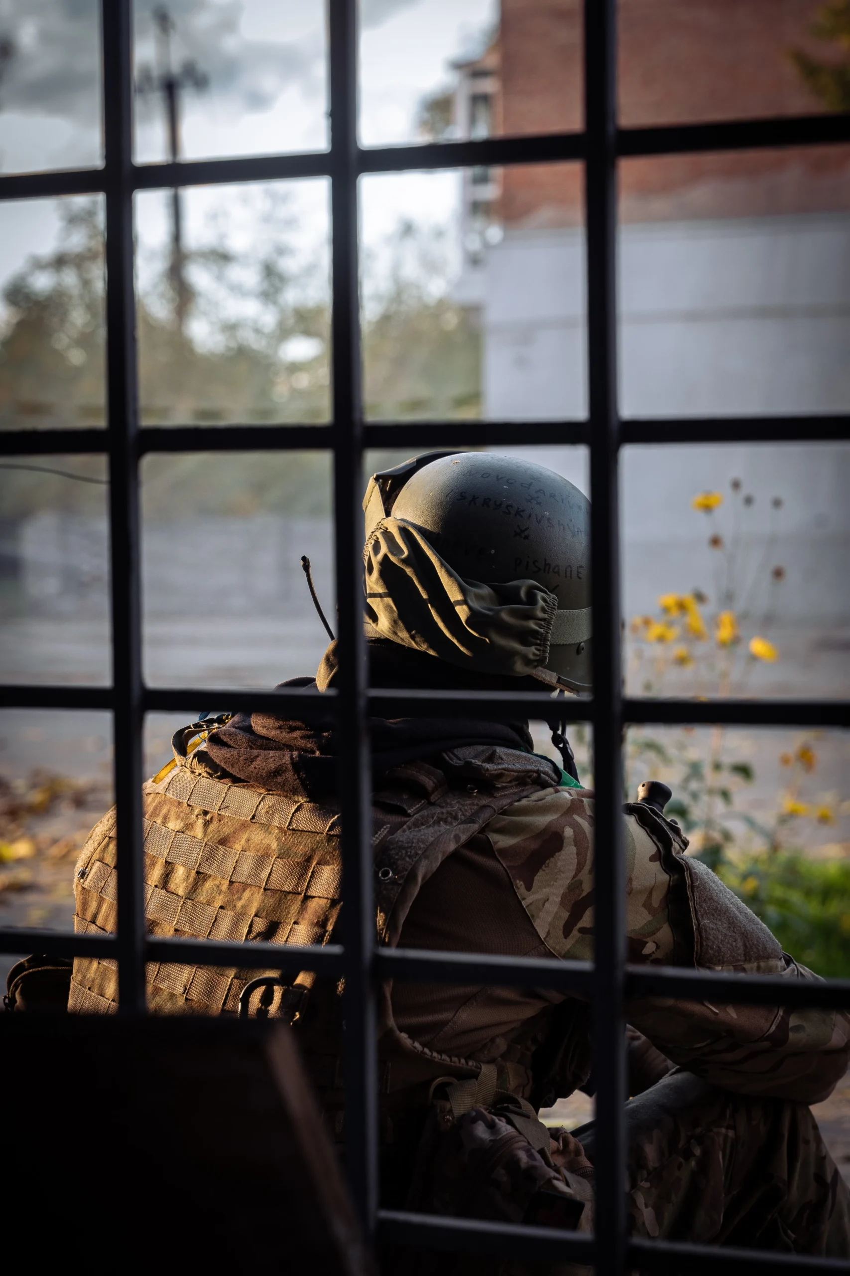 A soldier in a helmet and tactical vest seen from behind, looking out a window through a thick metal security grid.