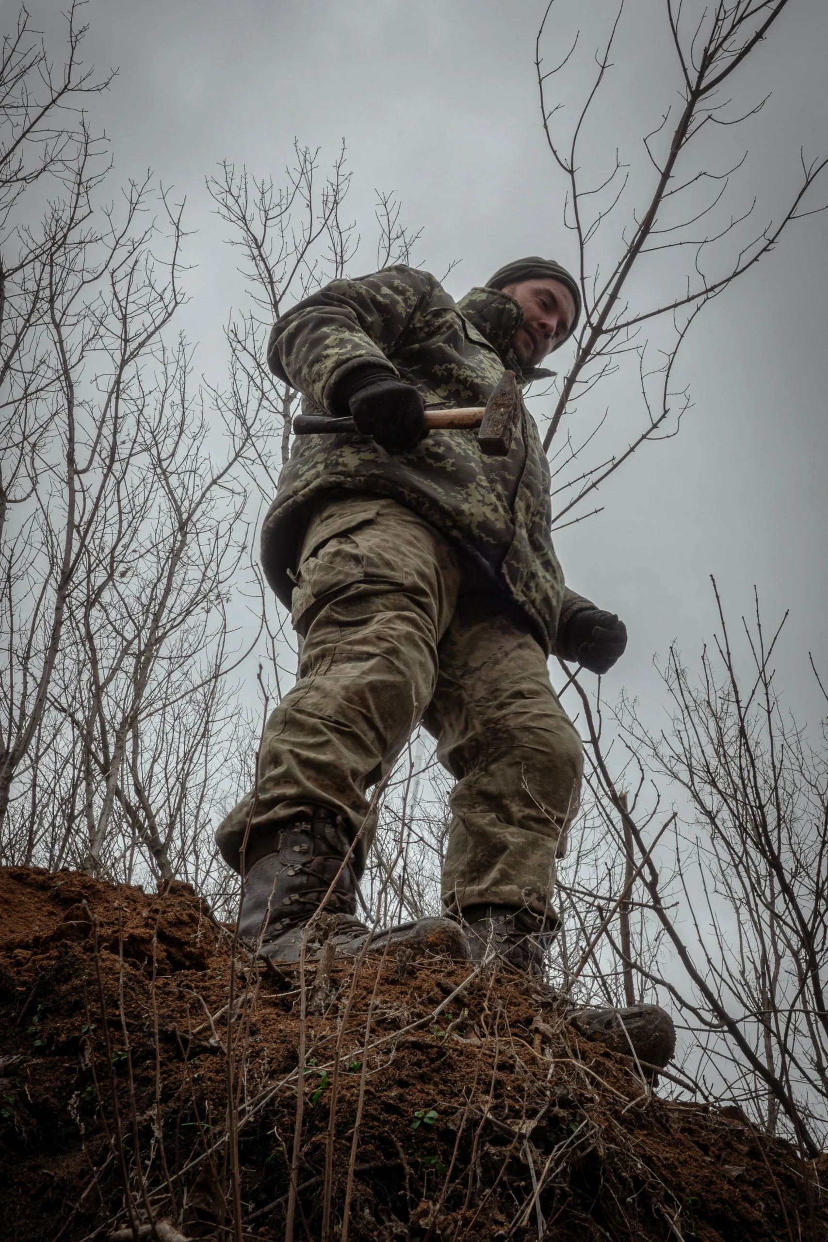 A low-angle shot of a soldier in camouflage standing on a dirt mound, holding a hammer against a cloudy grey sky.