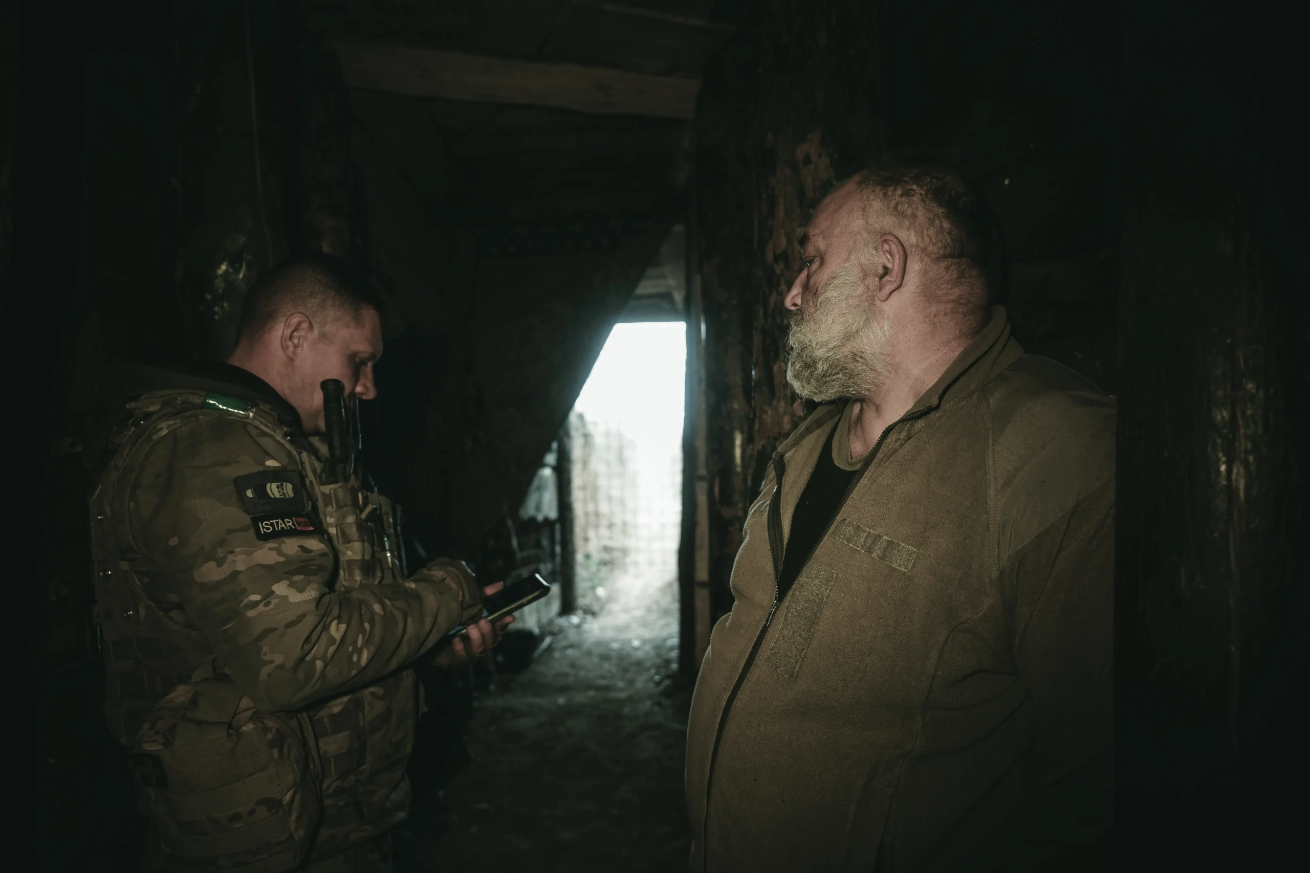 Two men standing in a dark, narrow underground bunker; one is checking a mobile device while the other looks on.